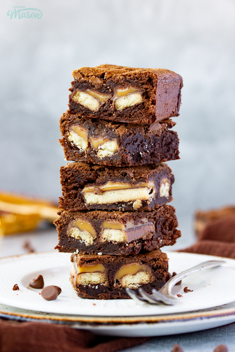 A stack of 5 Twix brownies on a white ribbed plate with a fork set over a grey mottled backdrop. There's a brown linen napkin, a Twix bar and chocolate chips scattered in the background.