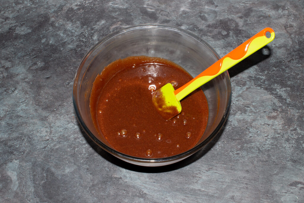 Melted butter, dark chocolate, eggs and sugar mixed together in a glass bowl with a green spatula.