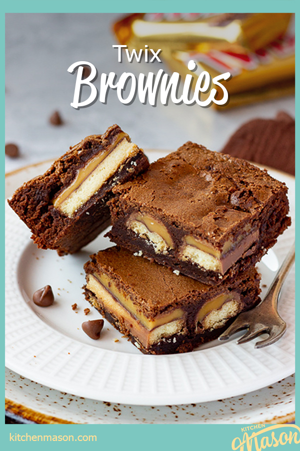 3 Twix brownies and a fork stacked on a white ribbed plate over a mottled grey backdrop. There are 2 Twix bars, a brown linen napkin and chocolate chips scattered in the background.