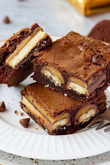 3 Twix brownies and a fork stacked on a white ribbed plate over a mottled grey backdrop. There are 2 Twix bars, a brown linen napkin and chocolate chips scattered in the background.