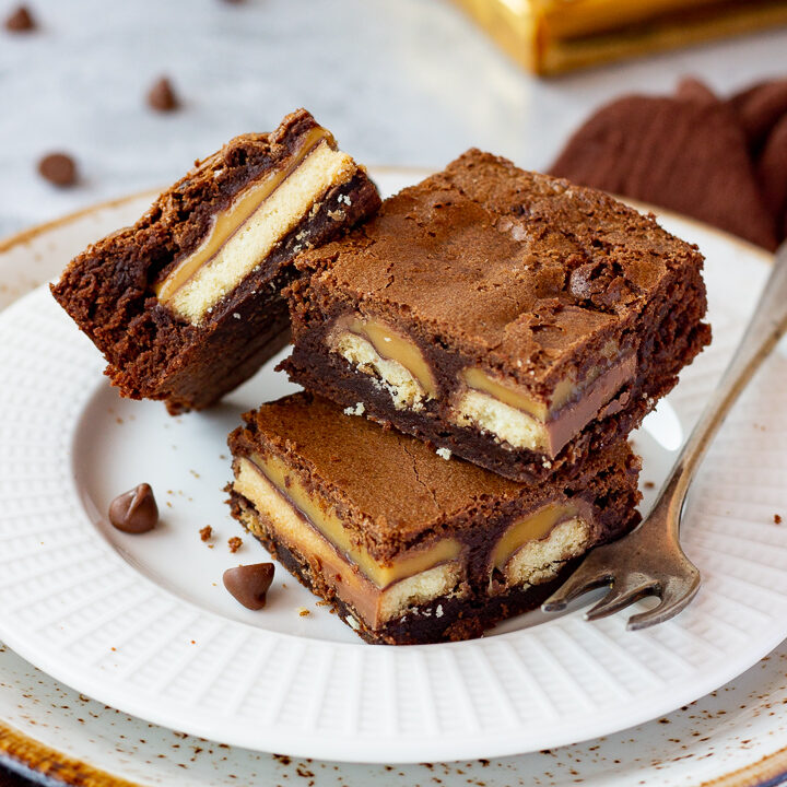 3 Twix brownies and a fork stacked on a white ribbed plate over a mottled grey backdrop. There are 2 Twix bars, a brown linen napkin and chocolate chips scattered in the background.