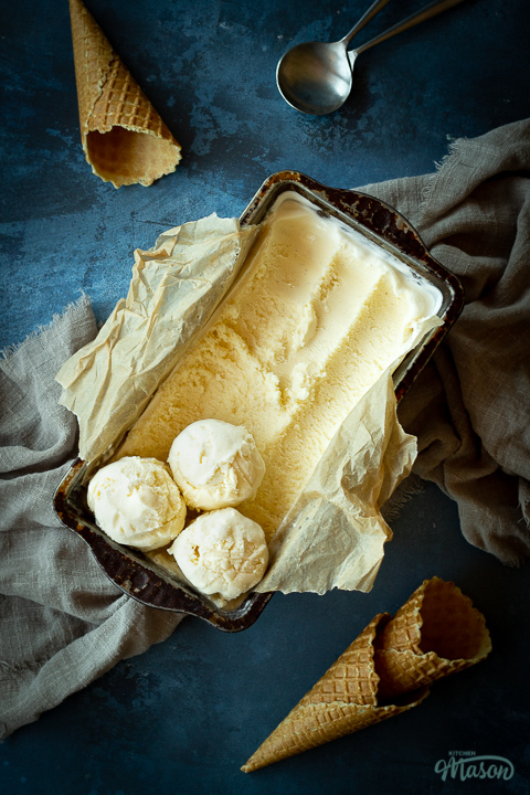 Vanilla ice cream in a lined loaf tin with 3 scoops at the end. Set over a crumpled light brown napkin over a deep blue mottled backdrop. There are 3 waffle cones and a spoon in the background.