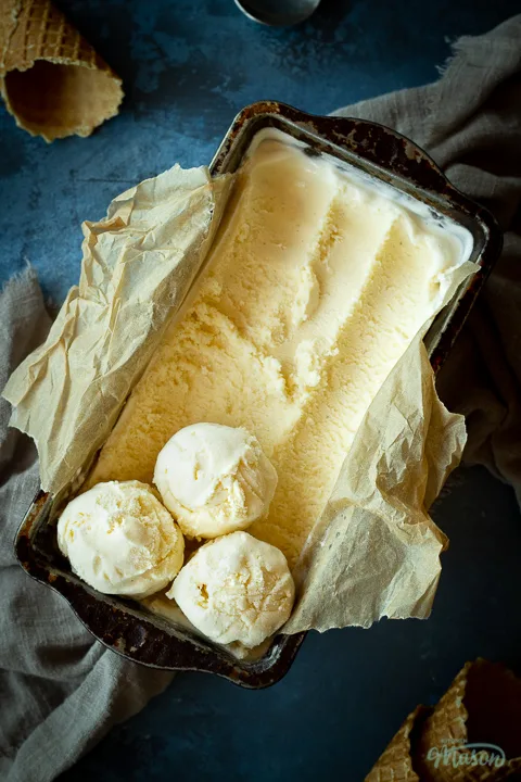 Close up of vanilla ice cream in a lined loaf tin with 3 scoops at the end. Set over a crumpled light brown napkin over a deep blue mottled backdrop. There are 3 waffle cones and a spoon in the background.