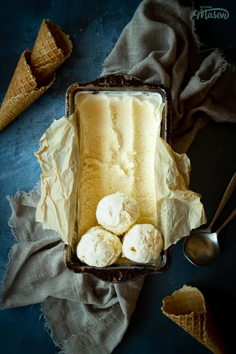 Vertical view of vanilla ice cream in a lined loaf tin with 3 scoops at the end. Set over a crumpled light brown napkin over a deep blue mottled backdrop. There are 3 waffle cones and a spoon in the background.