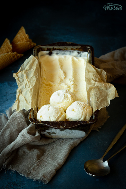 Front view of Vanilla ice cream in a lined loaf tin with 3 scoops at the end. Set over a crumpled light brown napkin over a deep blue mottled backdrop. There are 2 waffle cones and a spoon in the background.