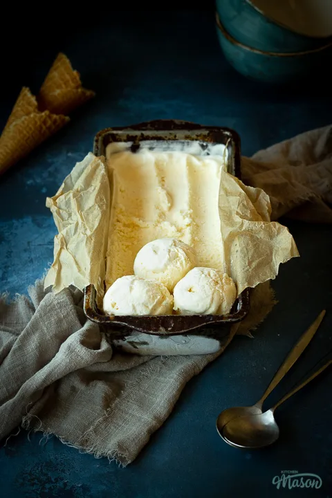 Front view of Vanilla ice cream in a lined loaf tin with 3 scoops at the end. Set over a crumpled light brown napkin over a deep blue mottled backdrop. There are 2 waffle cones and a spoon in the background.
