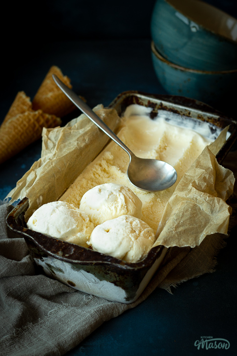 Diagonal side view of vanilla ice cream in a lined loaf tin with 3 scoops at the end and a spoon on top. Set over a crumpled light brown napkin over a deep blue mottled backdrop. There are 2 waffle cones and 2 stacked blue bowls n the background.