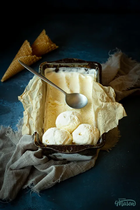 Font view of vanilla ice cream in a lined loaf tin with 3 scoops at the end and a spoon on top. Set over a crumpled light brown napkin over a deep blue mottled backdrop. There are 2 waffle cones in the background.