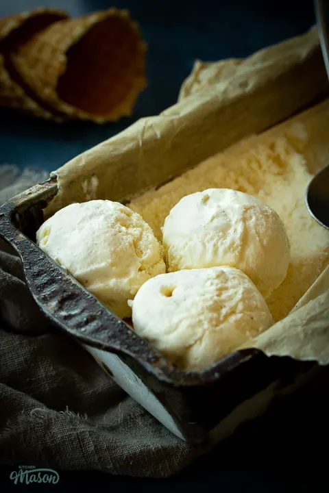 Close up side view of vanilla ice cream in a lined loaf tin with 3 scoops at the end and a spoon on top. There are 2 waffle cones in the background.