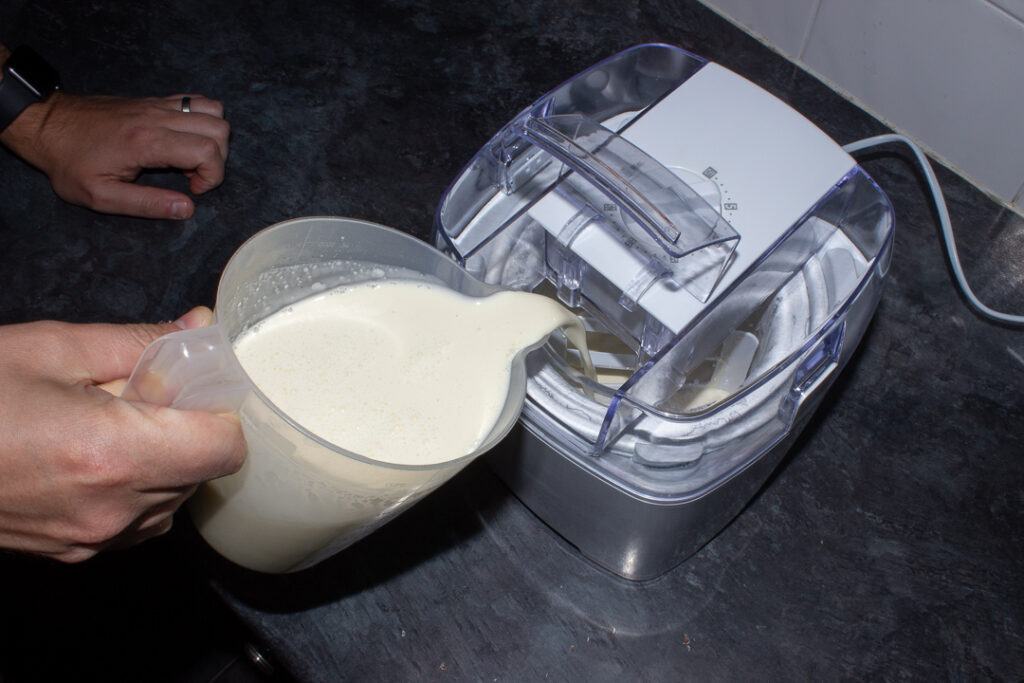 Vanilla ice cream mixture being poured into an ice cream churner on a kitchen worktop.