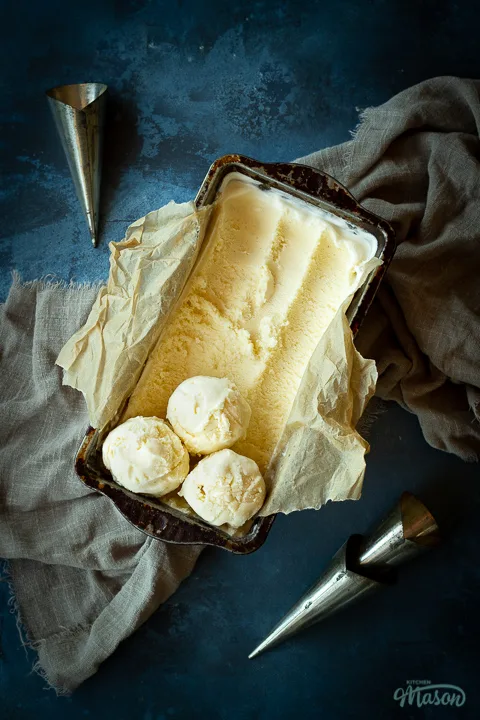 Vanilla ice cream in a lined loaf tin with 3 scoops at the end. Set over a crumpled light brown napkin over a deep blue mottled backdrop. There are 3 metal cone moulds in the background.