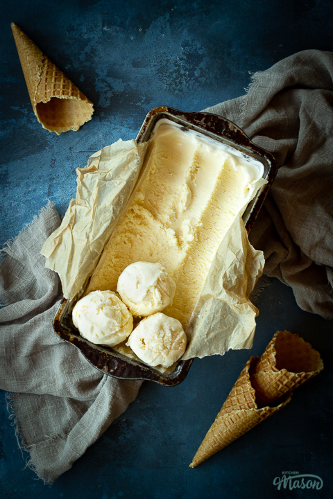 Vanilla ice cream in a lined loaf tin with 3 scoops at the end. Set over a crumpled light brown napkin over a deep blue mottled backdrop. There are 3 waffle cones in the background.