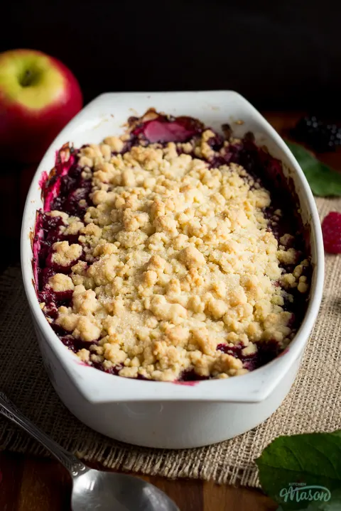 Front angled view of apple berry Summer fruit crumble on a piece of frayed hessian fabric over a wooden worktop. There are berries, leaves an apple and a spoon in the background.