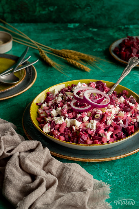 Side angle of apple and beetroot salad in a green serving dish on a dark green plate with a spoon inside. Set on a bright green backdrop with a light brown linen napkin, a plate of diced beetroot, some stacked green plates with cutlery on top, a pot of flaked almonds and some wheat in the background.