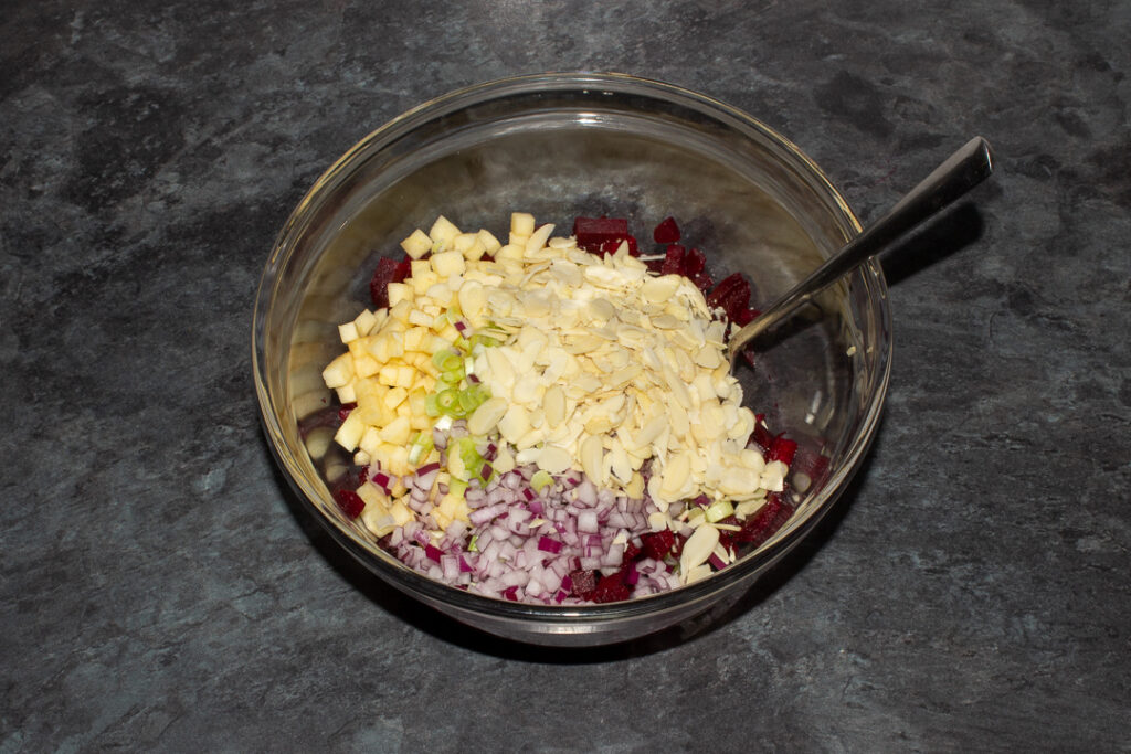 Diced beetroot, diced apple, spring onion, flaked almonds and red onion in a mixing bowl with a spoon on a kitchen worktop.
