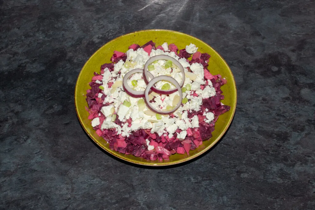 Apple and beetroot salad in a green serving bowl topped with creamy feta cheese, flaked almonds, spring onion and 3 rings of red onion.