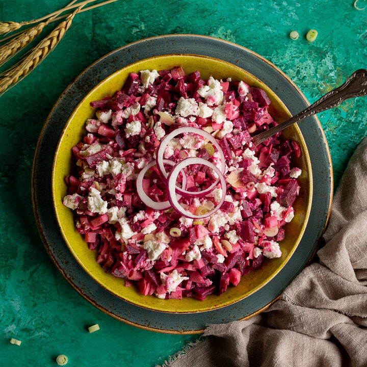 Flat lay view of apple and beetroot salad in a green serving dish on a dark green plate with a spoon inside. Set on a bright green backdrop with a light brown linen napkin, some wheat and chopped spring onion scattered in the background.