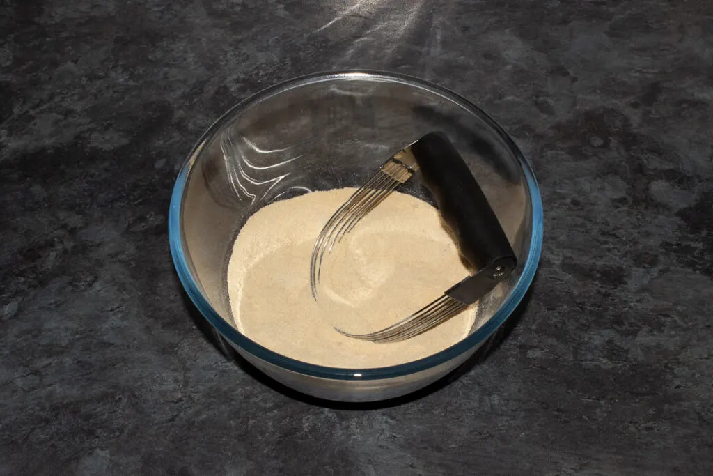 Flour, brown sugar and salt mixed together in a glass bowl with a pastry blender on a grey worktop.