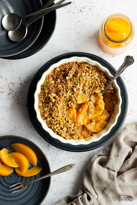 Peach crisp in a fluted ceramic serving dish set on a blue plate with a serving spoon inside. There's a light brown linen napkin, a blue plate of peaches with a fork, a jar of peaches and 2 stacked blue plates with spoons on top in the background. Set over a light mottled backdrop.