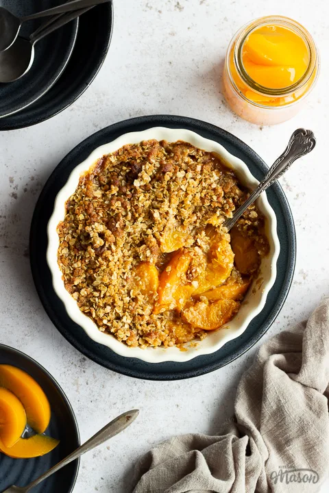 Peach crisp in a fluted ceramic serving dish set on a blue plate with a serving spoon inside. There's a light brown linen napkin, a blue plate of peaches with a fork, a jar of peaches and 2 stacked blue plates with spoons on top in the background. Set over a light mottled backdrop.