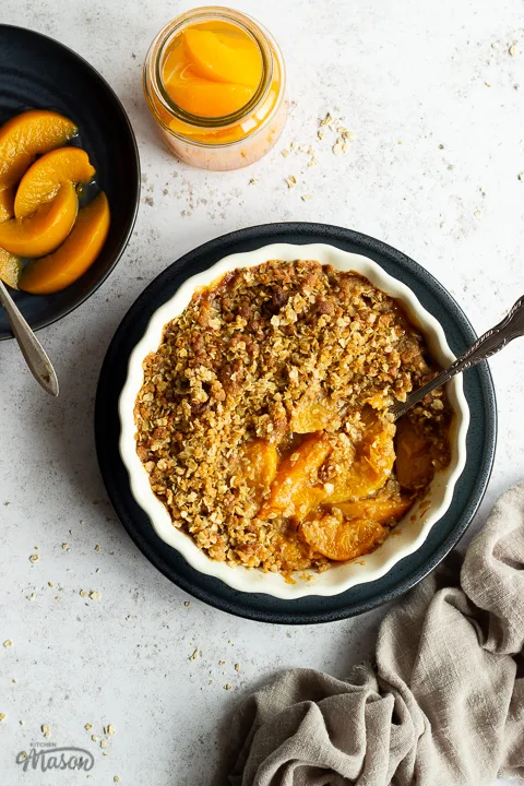 Peach crisp in a fluted ceramic serving dish set on a blue plate with a serving spoon inside. There's a light brown linen napkin, a blue plate of peaches with a fork and a jar of peaches in the background. Set over a light mottled backdrop.