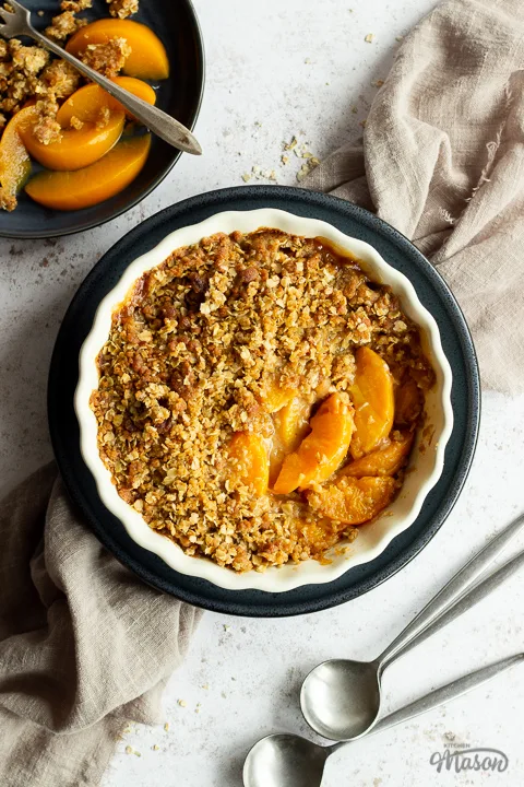 Peach crisp in a fluted ceramic serving dish set on a blue plate over a light brown linen napkin. There are two spoons and a blue plate with peach crisp on it in the background. Set over a light mottled backdrop.