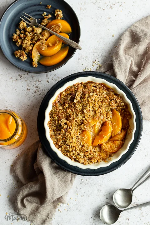 Peach crisp in a fluted ceramic serving dish set on a blue plate over a light brown linen napkin. There are two spoons, a blue plate with peach crisp on it and a jar of peaches in the background. Set over a light mottled backdrop.