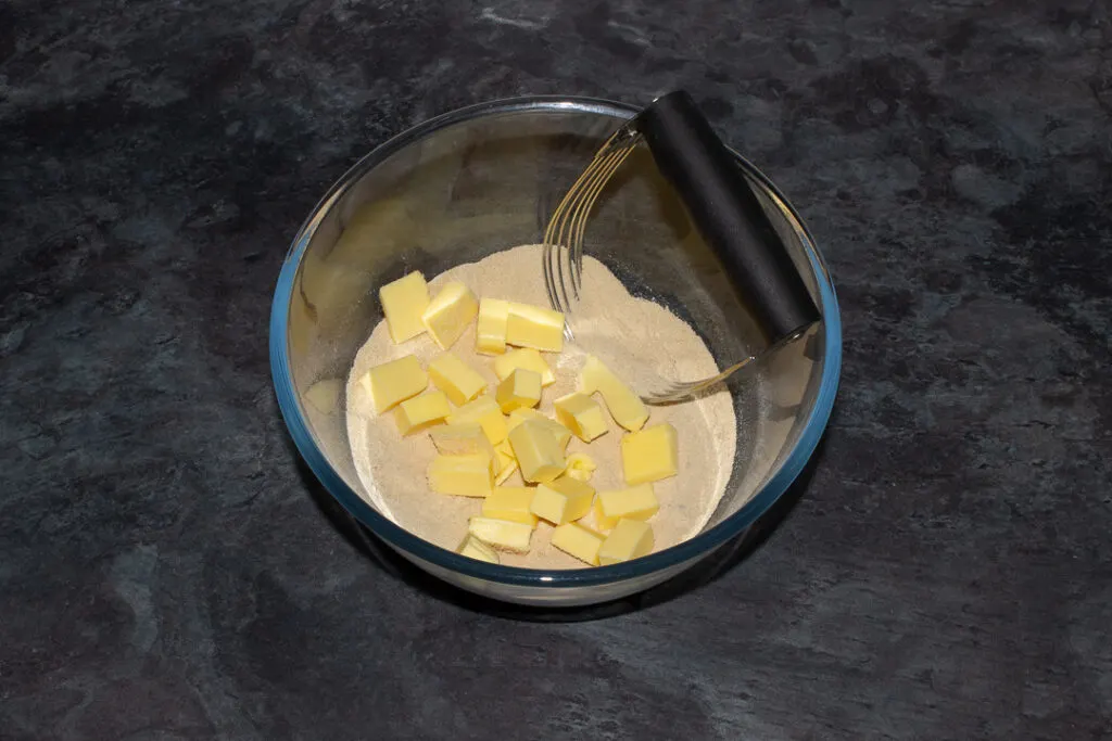 Flour, brown sugar and salt mixed together with cold cubed butter on top in a glass bowl with a pastry blender on a grey worktop.