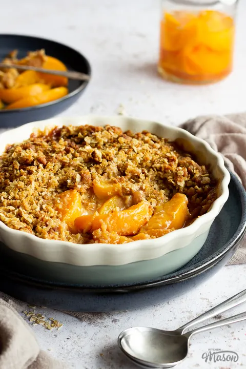 Side view of peach crisp in a fluted ceramic serving dish set on a blue plate. There's a light brown linen napkin, a blue plate of peaches with a fork, a jar of peaches and two spoons in the background. Set over a light mottled backdrop.