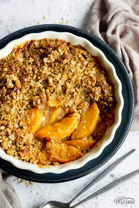 Close up of peach crisp in a fluted ceramic serving dish set on a blue plate over a light brown linen napkin. There are two spoons in the background and it's all set over a light mottled backdrop.