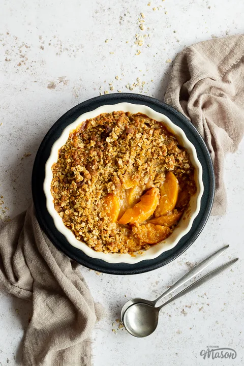 Peach crisp in a fluted ceramic serving dish set on a blue plate over a light brown linen napkin. There are two spoons in the background and it's all set over a light mottled backdrop.