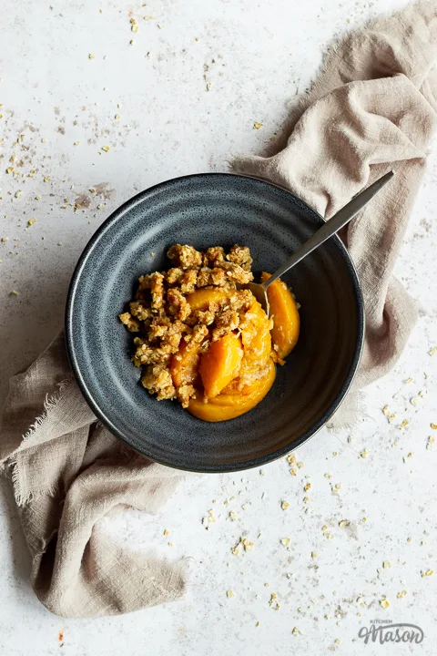 A blue bowl with peach crisp and a metal spoon inside set over a light brown linen napkin on a light mottled backdrop.