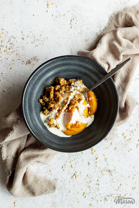 A blue bowl with peach crisp, some cream and a metal spoon inside set over a light brown linen napkin on a light mottled backdrop.