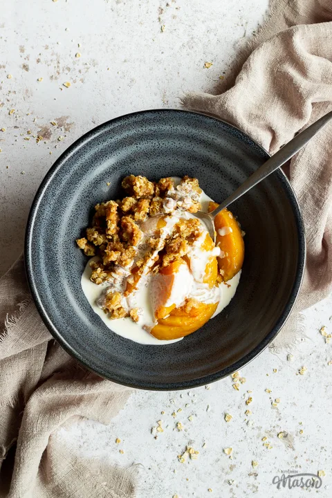 Close up of a blue bowl with peach crisp, some cream and a metal spoon inside set over a light brown linen napkin on a light mottled backdrop.