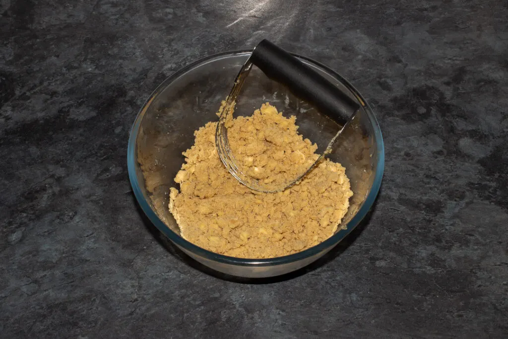 Flour, brown sugar, salt and butter mixed together in a glass bowl with a pastry blender on a grey worktop.