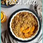 Peach crisp in a fluted ceramic serving dish set on a blue plate over a light brown linen napkin. There are two spoons, a blue plate with peach crisp on it and a jar of peaches in the background. Set over a light mottled backdrop.