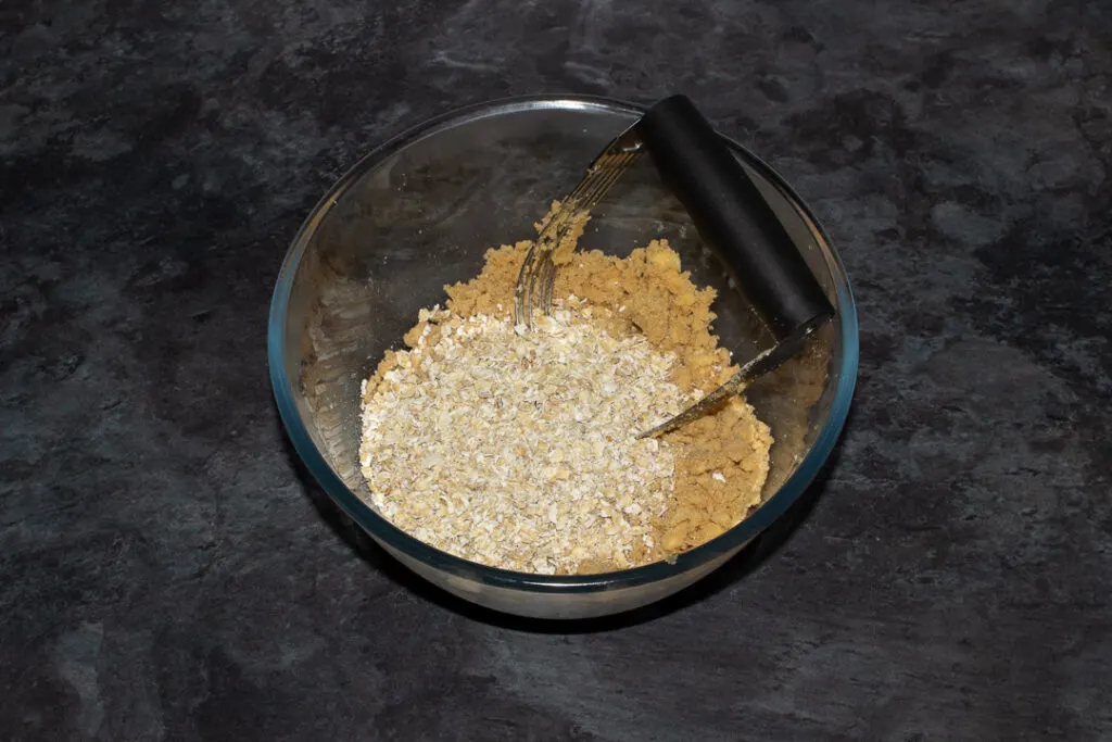 Flour, brown sugar, salt and butter mixed together with oats on top in a glass bowl with a pastry blender on a grey worktop.