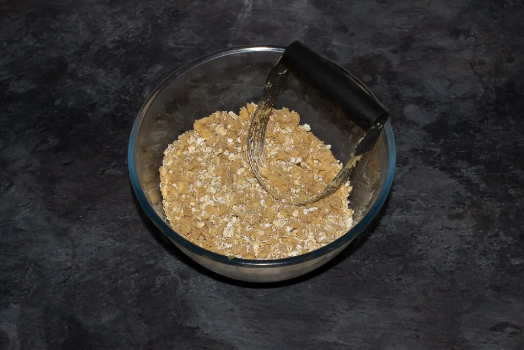 Flour, brown sugar, salt, butter and oats mixed together in a glass bowl with a pastry blender on a grey worktop.
