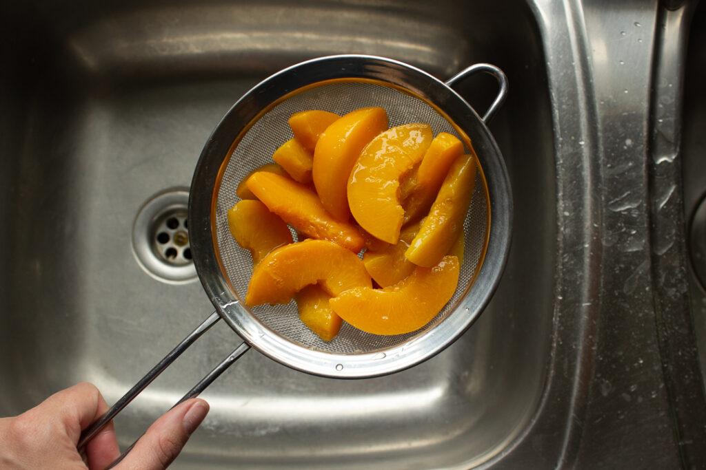 Tinned peaches being drained in a sieve set over a sink.
