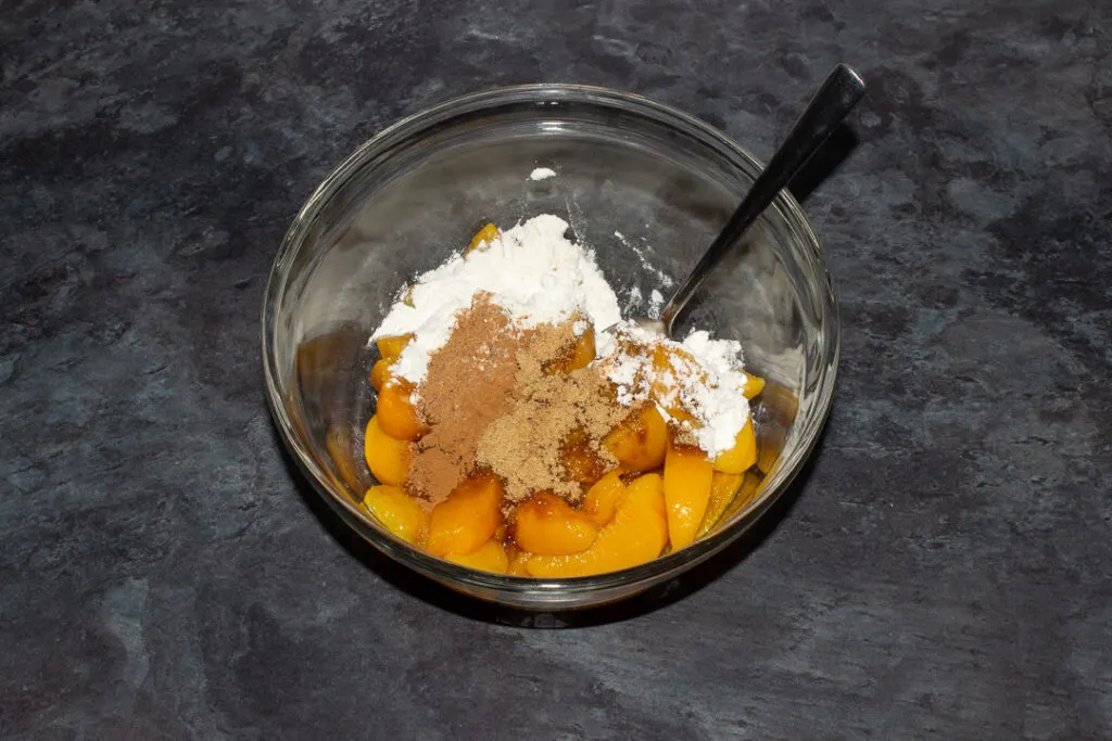 Drained tinned peaches, brown sugar, flour, nutmeg and cinnamon in a glass bowl with a metal spoon set on a grey worktop.
