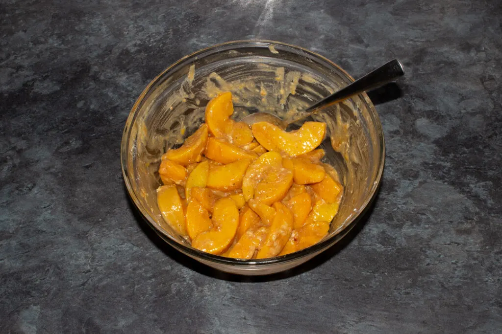 Drained tinned peaches, brown sugar, flour, nutmeg and cinnamon mixed together in a glass bowl with a metal spoon set on a grey worktop.