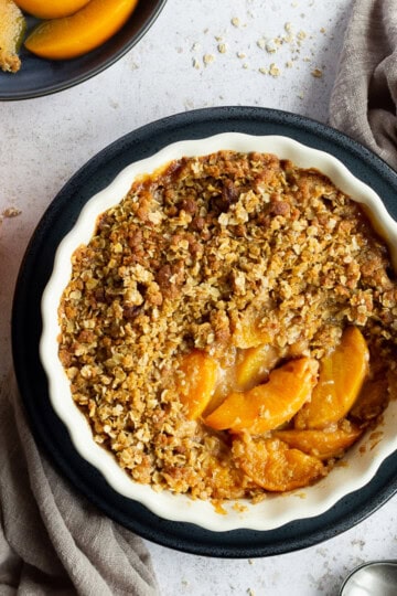 Peach crisp in a fluted ceramic serving dish set on a blue plate over a light brown linen napkin. There are two spoons, a blue plate with peach crisp on it and a jar of peaches in the background. Set over a light mottled backdrop.