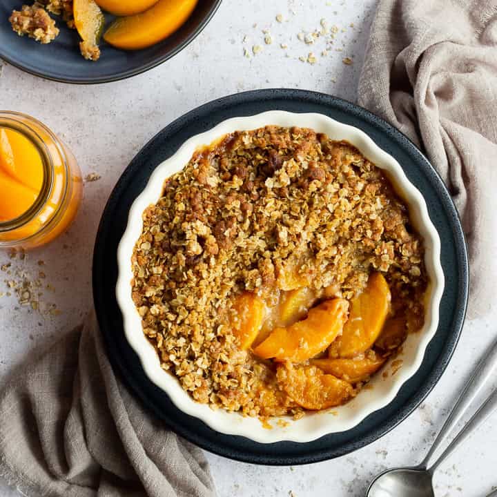 Peach crisp in a fluted ceramic serving dish set on a blue plate over a light brown linen napkin. There are two spoons, a blue plate with peach crisp on it and a jar of peaches in the background. Set over a light mottled backdrop.
