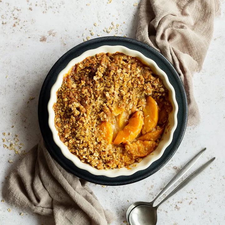 Peach crisp in a fluted ceramic serving dish set on a blue plate over a light brown linen napkin. There are two spoons in the background and it's all set over a light mottled backdrop.