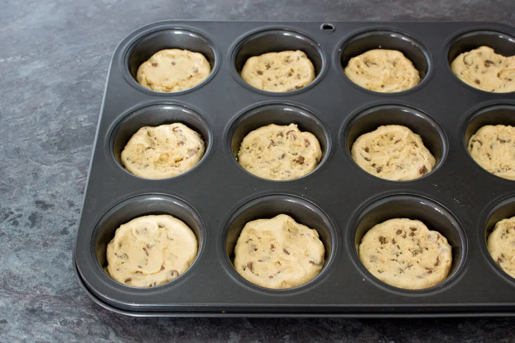 Cookie dough pressed into the holes of a cupcake/muffin tin on a kitchen worktop.
