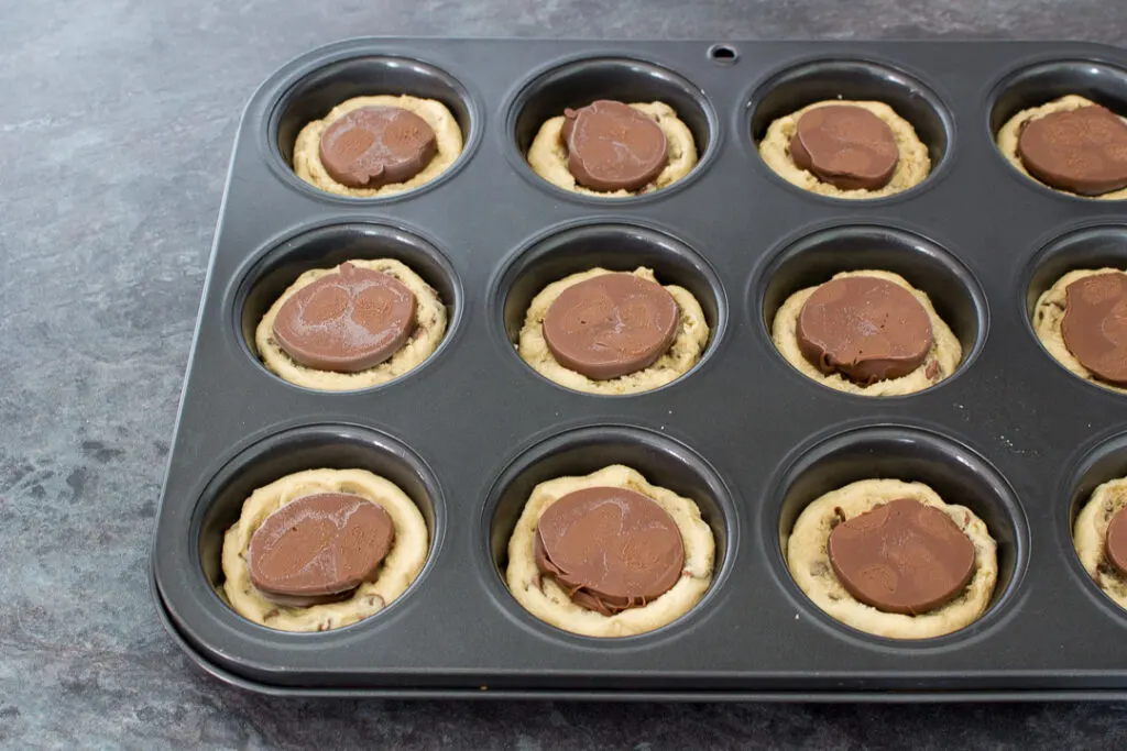 Cookie dough pressed into the holes of a cupcake/muffin tin with frozen Nutella blobs on top on a kitchen worktop.