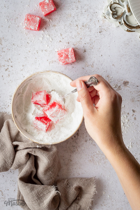 Close up of homemade Turkish delight in a bowl of icing sugar with a spoon being held by a hand. There's a light brown linen napkin resting against the bowl and several other pieces of Turkish delight scattered around the backdrop. Set on a white mottled backdrop.