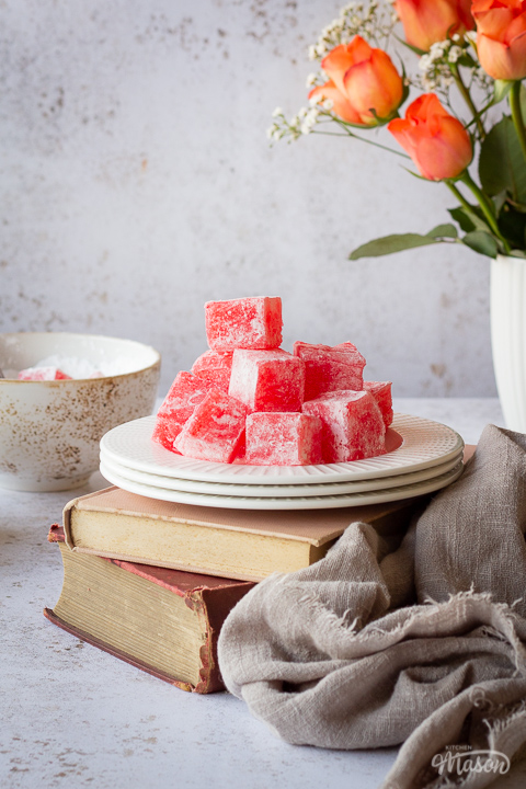 Close up view of a white plate filled with homemade Turkish delight set on top of two old books. There's a light brown linen napkin resting against the books and a bowl of icing sugar with a spoon and a white mug filled with orange roses in the background. All set on a white mottled backdrop.