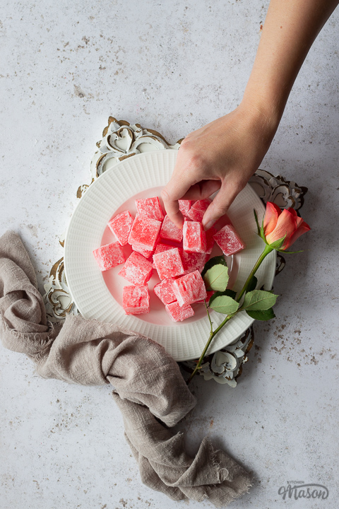 Homemade Turkish delight on a white plate with an orange rose set on a square rustic white board over a mottled white backdrop. There is a light brown linen napkin resting against the side and a hand reaching in to take a piece.