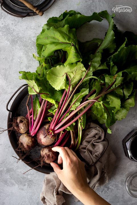 A bunch of whole beetroot complete with leaves covered in soil on a dark grey metal tray with a light brown linen napkin resting at the side and a hand holding it. There is also an open jar of pickled beetroot, an empty pickling jar and a couple of stacked grey metal plates with a wooden fork on top in the background. Set over a brushed grey backdrop.